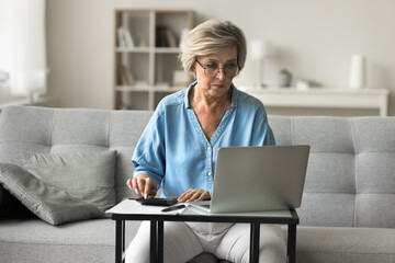 Serious older woman using laptop and calculator to control finances, pay household bills, taxes online via secure e-bank system, review account balance and recent transactions. Financial management