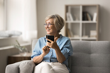 Attractive smiling elderly woman sitting on couch, holding smartphone, look away, connecting with...