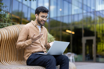 Young man wearing earphones using laptop outdoors sitting on wooden bench. Engaging in video call showing excitement and connectedness. Modern building setting enhances business and communication