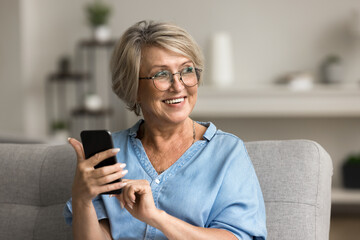 Pretty middle-aged woman in glasses looking away holding smartphone smile, resting on sofa at home, making call, browse wireless internet, search information, spend free time online on leisure indoors