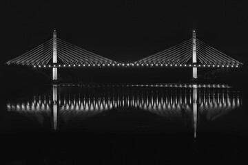 Side view of a suspender cables bridge over a river at night, with its reflection on the water. B&W image.