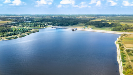 A scenic aerial view of a reservoir with a dam, surrounded by lush green fields and forests. The calm water reflects the sky, and the countryside stretches towards the horizon under a clear sky.