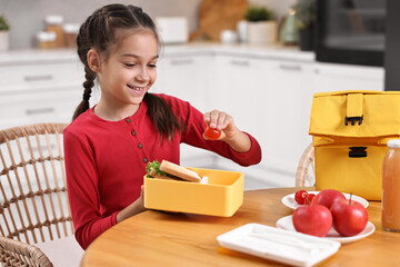 Cute girl putting tomato into lunch box at wooden table in kitchen. Preparing for school