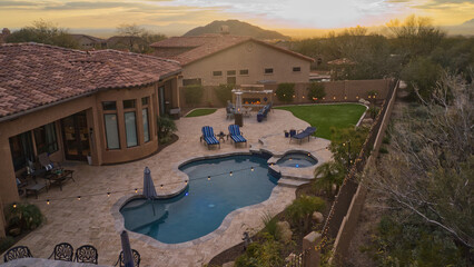 An aerial view of a desert landscaped backyard featuring a travertine pool deck with an outdoor kitchen, fireplace, pool and spa.