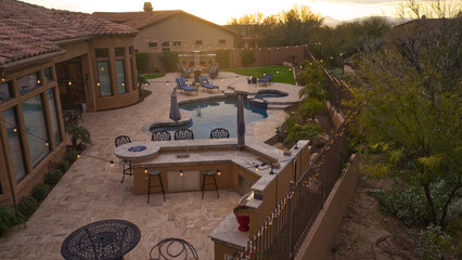 An aerial view of a desert landscaped backyard featuring a travertine pool deck with an outdoor kitchen, fireplace, pool and spa.