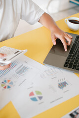Close-up of a person working on a laptop and calculator with financial charts on a yellow desk,...