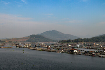 Fototapeta premium A stunning waterscape featuring fish farms floating on a lake, framed by distant green hills and mountains under a bright blue sky. A peaceful rural aquatic landscape