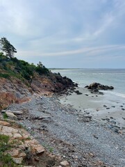 A coastal road lined with large rocks, facing a vast ocean under a cloudy sky. The waves gently crash, creating a peaceful seascape. Maine. Coastal and travel-themed decor.