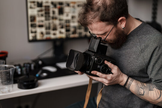 A bearded photographer with a tattoo carefully examines a vintage camera in a well-equipped photography studio with an artistic background.