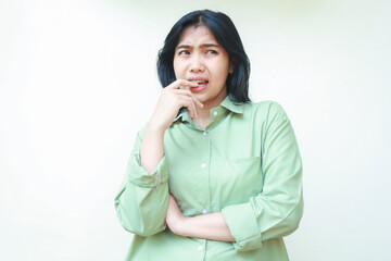 thoughtful asian woman with dark hair bite her nails and looking away curious with folding arms wearing green oversized shirt, annoyed female feeling afraid and scared, isolated on white