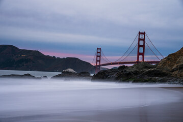 Golden Gate Bridge at dusk with misty waves and rocky shore.