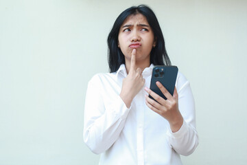 Fototapeta premium elegant asian young business woman daydreaming using smartphone wearing white formal shirt with finger on chin looking up confused what to buy in online shopping sale standing over isolated background