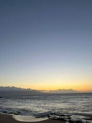 A Maui sunset paints the sky in blue, orange, and yellow. The calm waves reflect the warm colours, while clouds drift and a faint moon glows softly, completing the serene beach scene.
