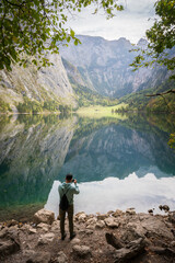 Vertical shot of man taking photo on bank of still beautiful greenish alpine lake, Obersee, Germany