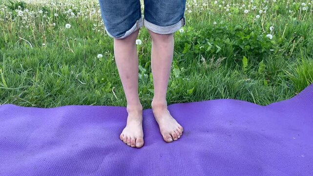 Child slowly transfers body weight from one foot to the other while standing barefoot on a purple yoga mat in the grass, dirty feets