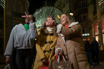 Three friends shopping at night under Christmas lights, pointing and smiling