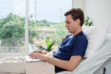 Side view of patient on hospital bed typing on laptop, combining recovery and productivity, representing resilience and modern digital healthcare environment.