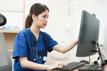 Portrait of smiling Southeast Asian nurse in blue uniform sitting at computer desk in bright modern...