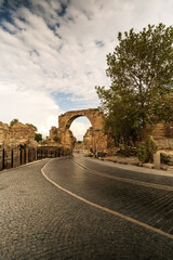 Fototapeta premium A winding cobblestone road leads towards a majestic ancient archway in the Side ruins, Turkey. Lush greenery frames the scene against a dramatic sky.