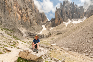Caucasian hiker with hat posing on a big rock at the Sassolungo (Langkofel) during summer. Hiking trail in Italian Dolomites during day time. Wonderful hiking area in mountains. World heritage site.
