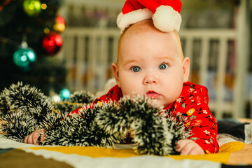 Newborn baby boy in a red Santa hat lies on the bed near the Christmas tree