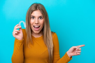 Young caucasian woman holding invisible braces isolated on blue background surprised and pointing finger to the side