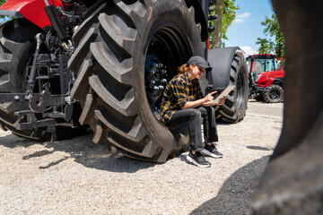 Female farmer sits on the big wheel of farm tractor and using her digital tablet.