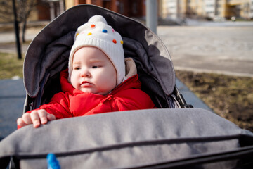 Baby in a Red Jacket and White Hat Seated in a Stroller on a Sunny Day. A baby dressed warmly looks on while sitting comfortably in a stroller.