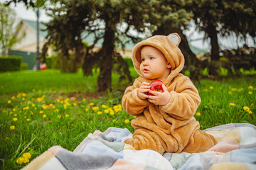 Cute baby girl in plush suit with red apple in hands sitting on blanket in green grass.