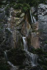 Obraz premium A multi-tiered waterfall cascades over rugged, moss-covered rocks in a forested area. The water flows gracefully through the steep rock formations, creating a serene and picturesque natural scene.