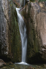 A narrow waterfall gracefully flows down between moss-covered rocks into a clear pool below. The contrast between the soft water and rugged rocks creates a serene and peaceful natural scene.
