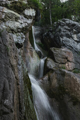 A cascading waterfall flows down steep, mossy rocks in a lush forest. The layers of rocks create a stunning visual as the water moves gracefully, carving its way through the rugged landscape.
