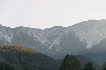 A serene view of majestic mountains, with their rocky peaks towering above dense forests. The sunlight gently illuminates the treetops, creating a beautiful contrast between the dark greenery and the 