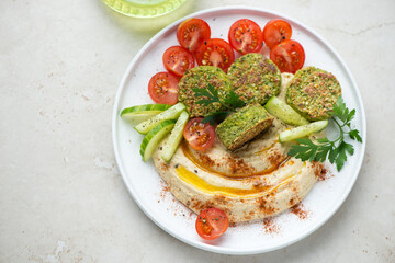 Hummus with paprika, falafel and fresh vegetables on a white plate, horizontal shot on a light-beige stone background, high angle view