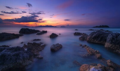 Obraz premium The sky transitions to deep hues as the sun sets, casting reflections on the calm water near the rocky coast