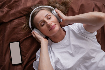 Woman in headphones lying on bed with eyes closed, smartphone resting beside her, enjoying peaceful...