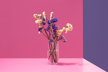 Ramo de flores secas azules y amarillas en un jarrón con agua sobre un fondo rosa	