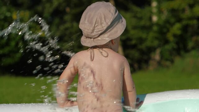 Infant child in stylish sun panama hat standing backside in inflatable pool, embodying summer and creating unforgettable memories. Baby boy stands with his back, splashing in backyard, slow motion
