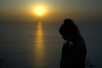 Fototapeta premium Silhouette of a woman at sunset by the sea. Hands folded in prayer, head down. The day is ending, the sky is painted in warm tones by the rays of the setting sun and leaves its mark on the sea.