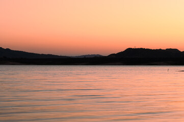 Fototapeta premium Soft pink tones of sunset reflect on lake, with silhouettes of hills creating stunning backdrop