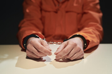 Person's hands handcuffed on table in dimly lit room, wearing orange jumpsuit suggesting...
