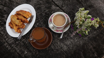 Tea, coffee and snacks (fried bananas) are served on a wooden table, with a natural landscape in the background.