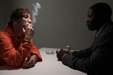 Two men sitting across from each other discussing legal matters in a prison meeting room, one wearing an orange jumpsuit and the other in a suit, smoke coming from cigarette visible