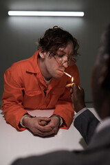 Man in orange jumpsuit leaning on table while another person lights his cigarette in dimly lit...