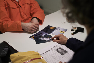 Two people discussing evidence photos in investigation room, one person wearing orange jumpsuit and another person holding criminal profile