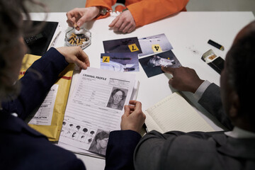Investigators examining criminal profiles and evidence at desk filled with photos, papers, and...