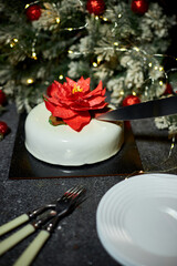 A person is cutting a festive cake adorned with a red flower, surrounded by christmas holiday decorations, glowing lights, and a joyful atmosphere perfect for winter celebrations.