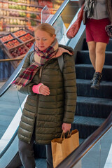 A woman with a pink cast on her right arm wearing a coat walks down an escalator at a supermarket,...