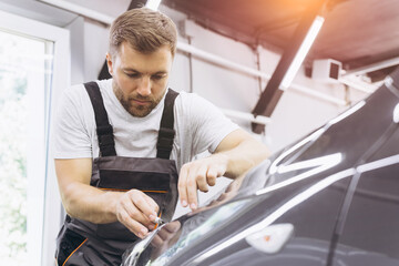 Detailing film paint protection, male specialist cutting and wrapping car in garage showroom,...