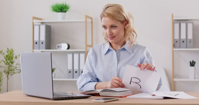 Speech therapist or foreign language teacher gives online lesson. Friendly woman at table with laptop waves hand, says hello, greets student, shows sheet of paper and explains how to pronounce sound R